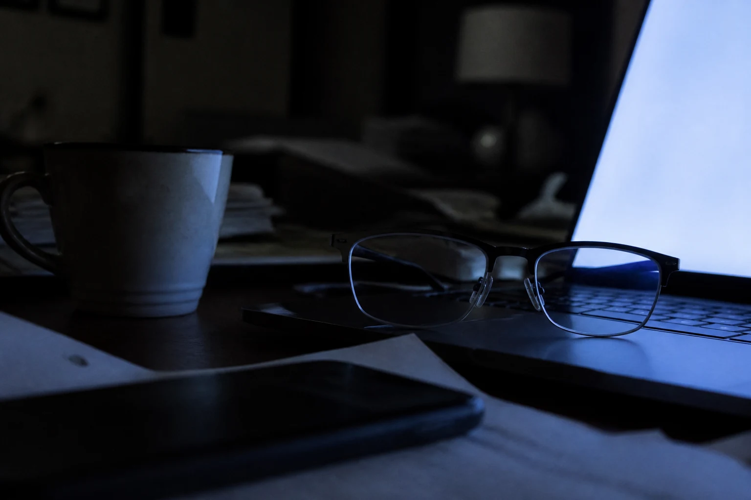 Egy szemüveg a billentyűzet mellett az asztalon, késő este, kizárólag a laptop képernyőjének kék fénye világítja meg a szobát, a lencsékben tükröződik a fény.